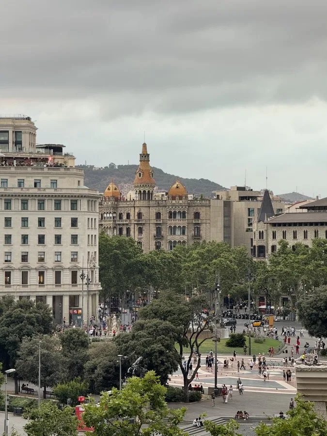 A square in Barcelona, a stone building with golden domes and a hill behind the trees